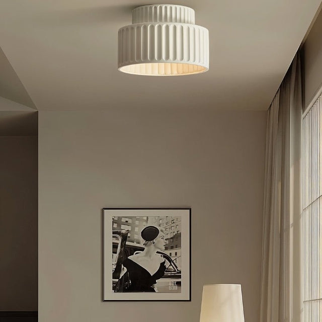 "A white ceiling light with a ribbed, cylindrical shade, shown installed on a white ceiling in a living room with a framed picture and a table lamp."