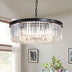 Black metal drum chandelier with clear crystal prisms hanging in a bright living room above a shelf with plants and books.
