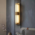 Brass and alabaster vertical wall sconce with warm light mounted between two dark textured walls in a modern bedroom.