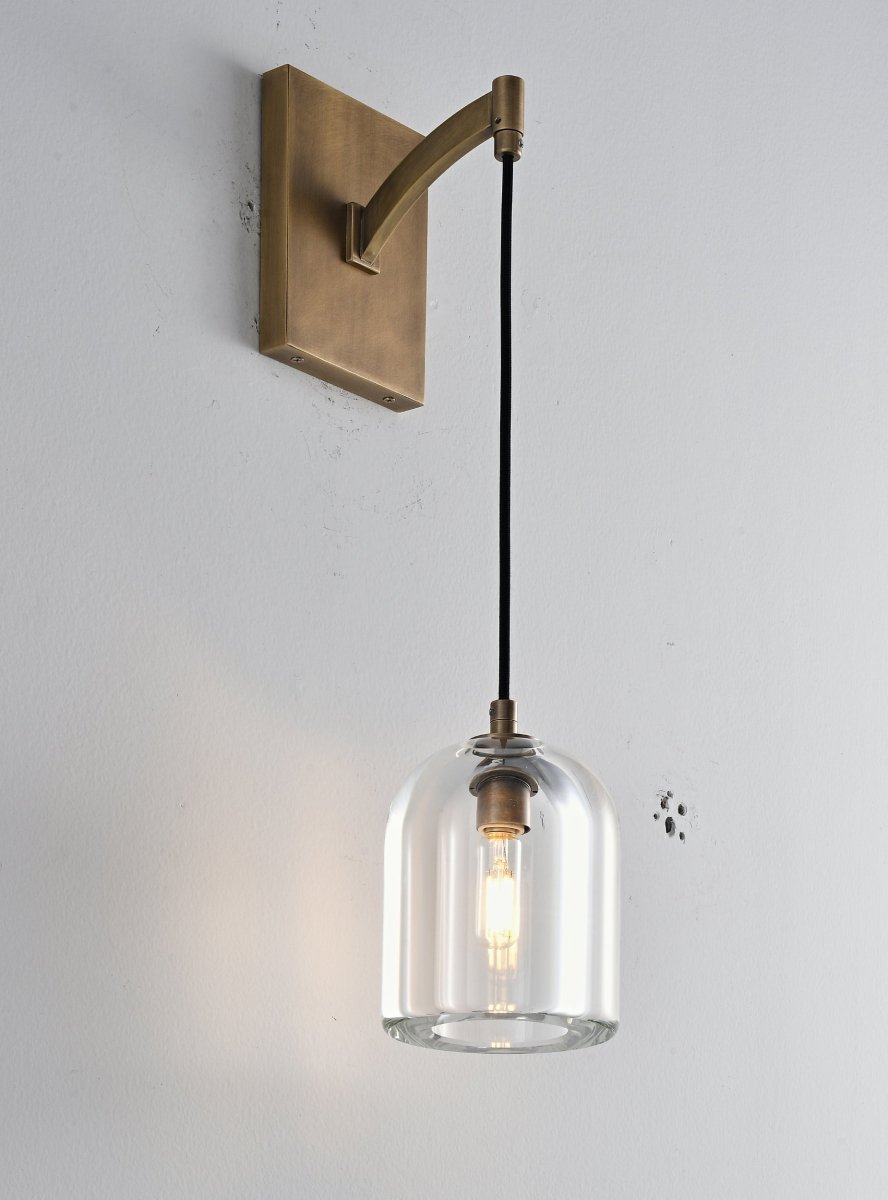 "A modern brass wall lamp with a bell-shaped clear glass shade, blending elegance and minimalist design with a curved arm and rectangular base."