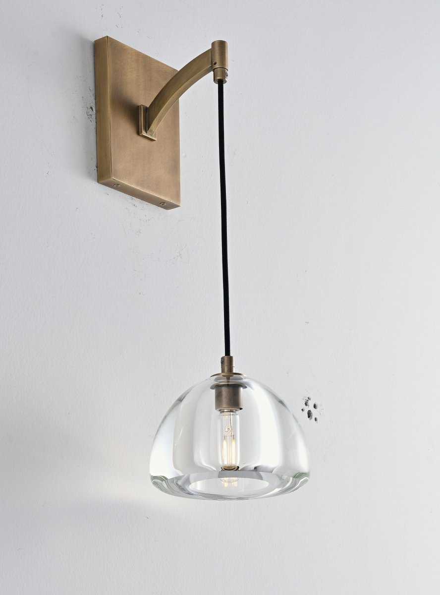 "A modern brass wall lamp with a dome-shaped clear glass shade, blending elegance and minimalist design with a curved arm and rectangular base."