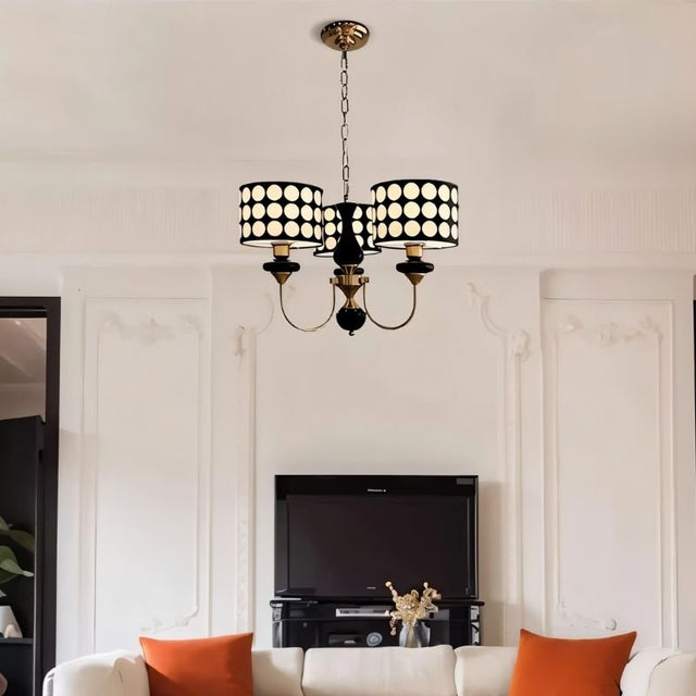 "A three-light chandelier with black polka dot shades hangs in a living room with a television and sofa."