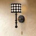 "A walnut and gold wall lamp with a patterned black shade and pull chain, installed in a bedroom with a curtain and a table."