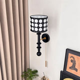 "A black and gold wall lamp with a patterned black and white shade and pull chain, viewed from above against a white wall."