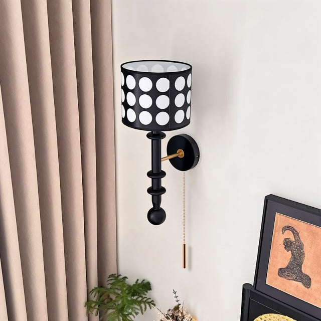 "A black and gold wall lamp with a patterned black and white shade and pull chain, viewed from above against a white wall."