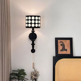 "A black and gold wall lamp with a patterned black shade and pull chain, installed on a wall with a picture frame and a plant."