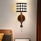 "A walnut and gold wall lamp with a patterned black shade and pull chain, installed in a bedroom with a wooden bed frame and a curtain.