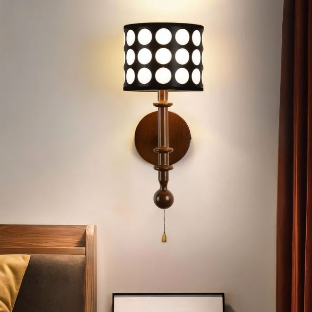 "A walnut and gold wall lamp with a patterned black shade and pull chain, installed in a bedroom with a wooden bed frame and a curtain.