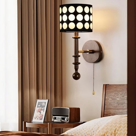 "A walnut and gold wall lamp with a patterned black shade and pull chain, installed in a bedroom with a curtain and a table."