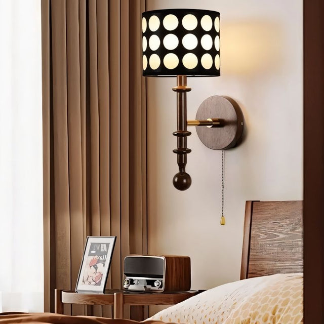 "A walnut and gold wall lamp with a patterned black shade and pull chain, installed in a bedroom with a curtain and a table."
