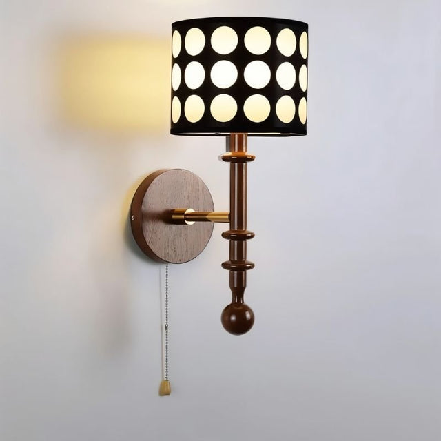 "A walnut and gold wall lamp with a patterned black shade and pull chain, lit up on a white wall."